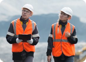 Two occupational health workers in high vis jackets and safety helmets walking outdoors and chatting at a mining site