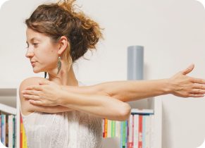Woman wearing a white sleeveless top stood in front of a bookcase, holding an occupational health arm stretch.